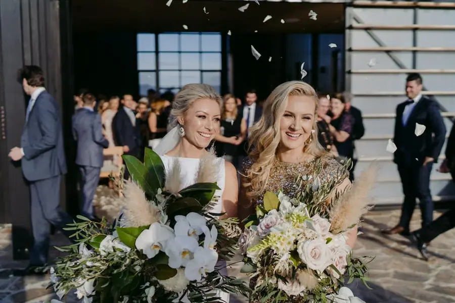 Two brides smiling with their bouquets as guests throw confetti outside the ceremony building