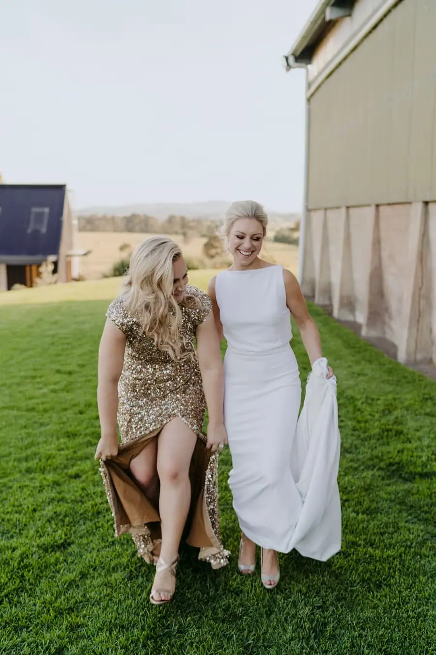 Two brides walking arm in arm across grass while lifting their dresses