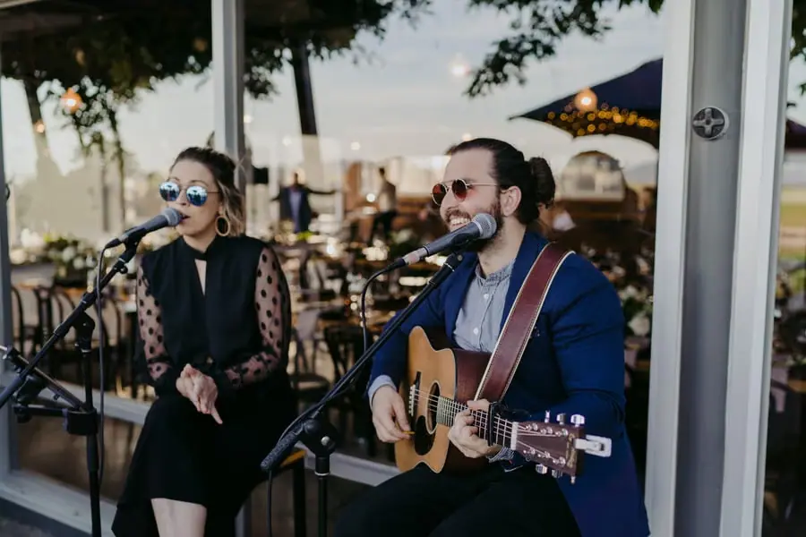 Two musicians singing into microphones with an acoustic guitar outdoors