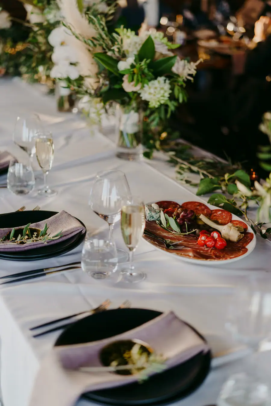Reception place settings and a platter of tomatoes beside white floral arrangements