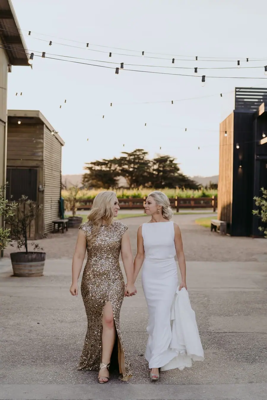 Two brides walking together beneath festoon lights between venue buildings