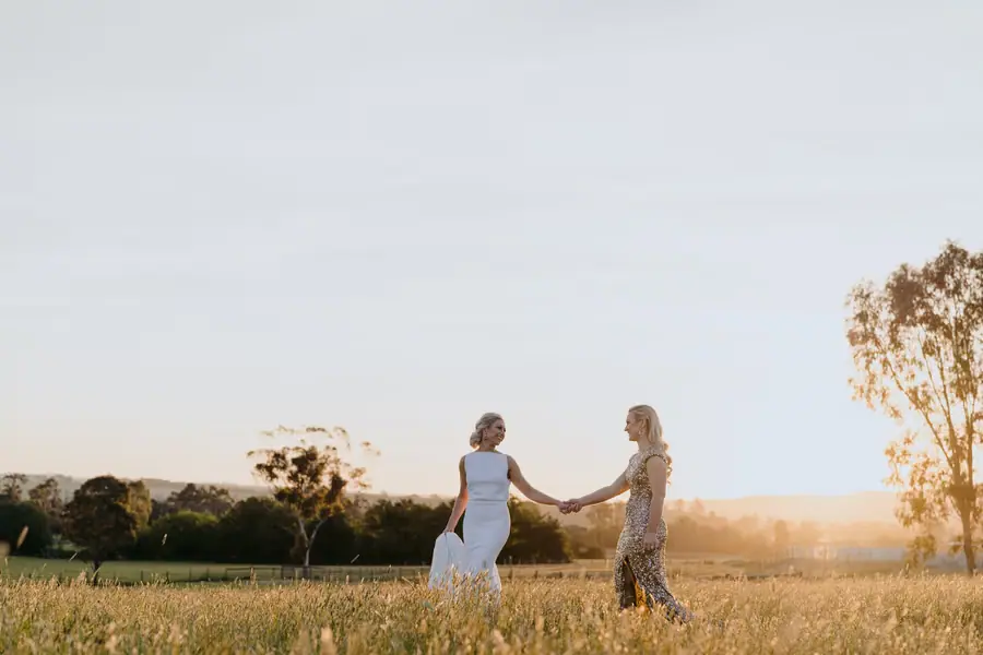 Two brides holding hands across a grassy field at sunset