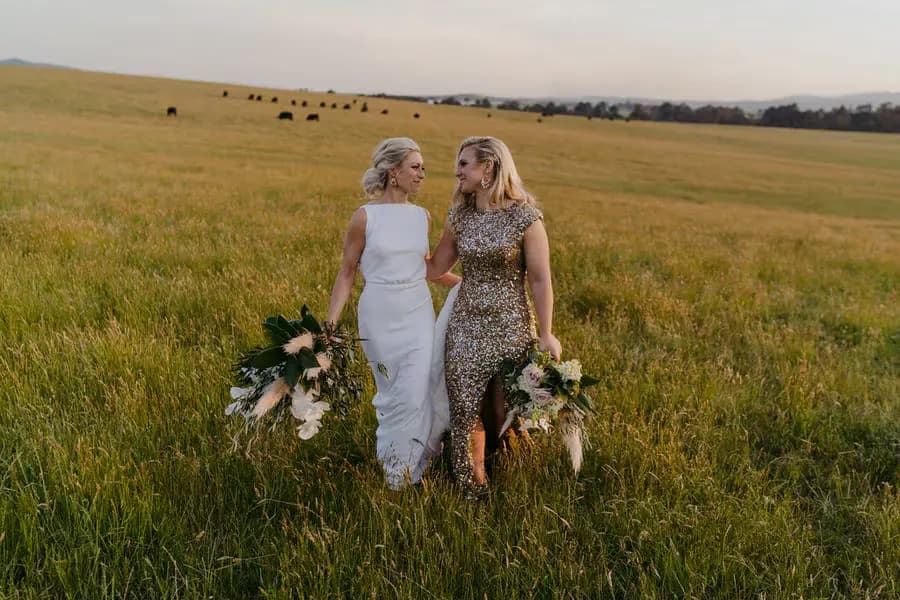 Two brides walking hand in hand through long grass with grazing animals behind