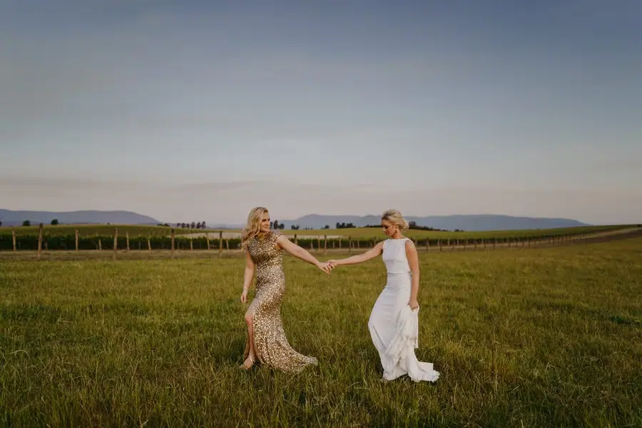 Two brides holding hands and turning toward one another beside vineyard rows