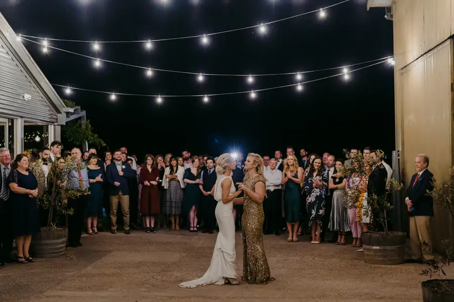 Two brides embracing beneath outdoor festoon lights as guests watch