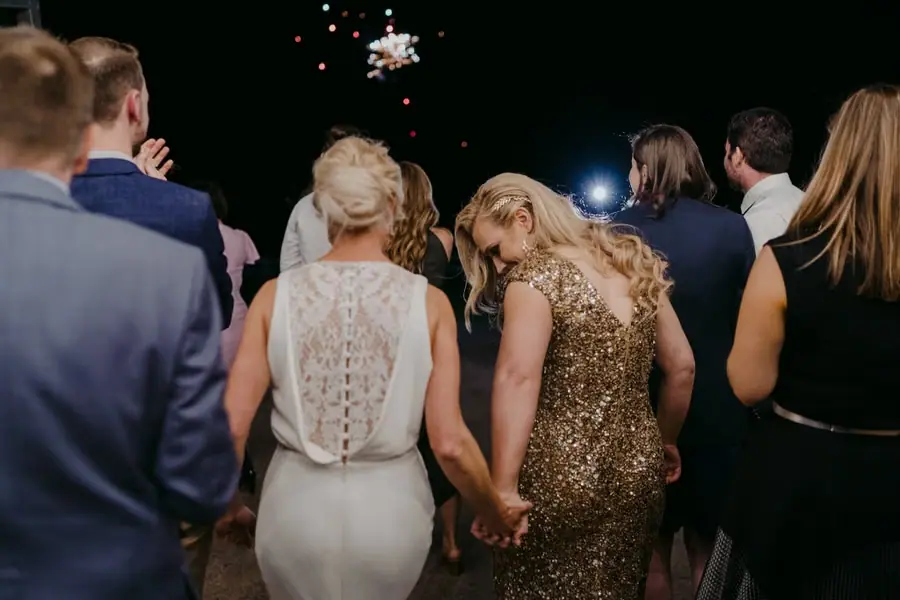 Two brides holding hands among guests watching fireworks in the night sky