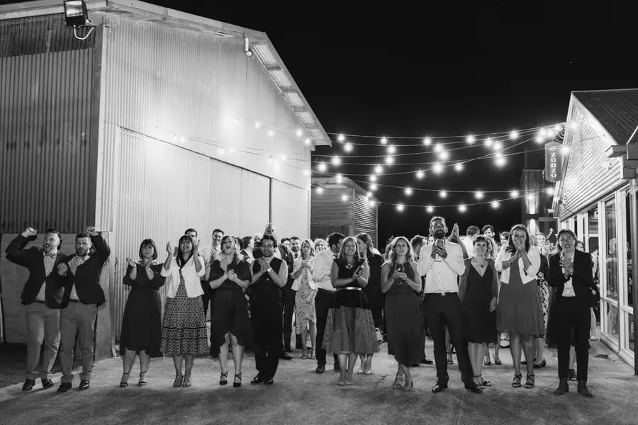 Black-and-white photograph of guests applauding beneath outdoor festoon lights