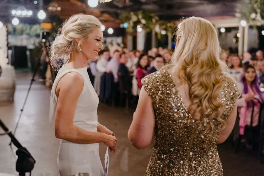 Two brides smiling toward one another near a microphone inside the reception