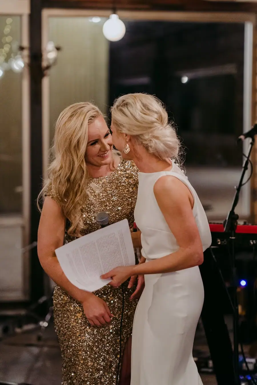 Two brides leaning close together while holding papers near a microphone