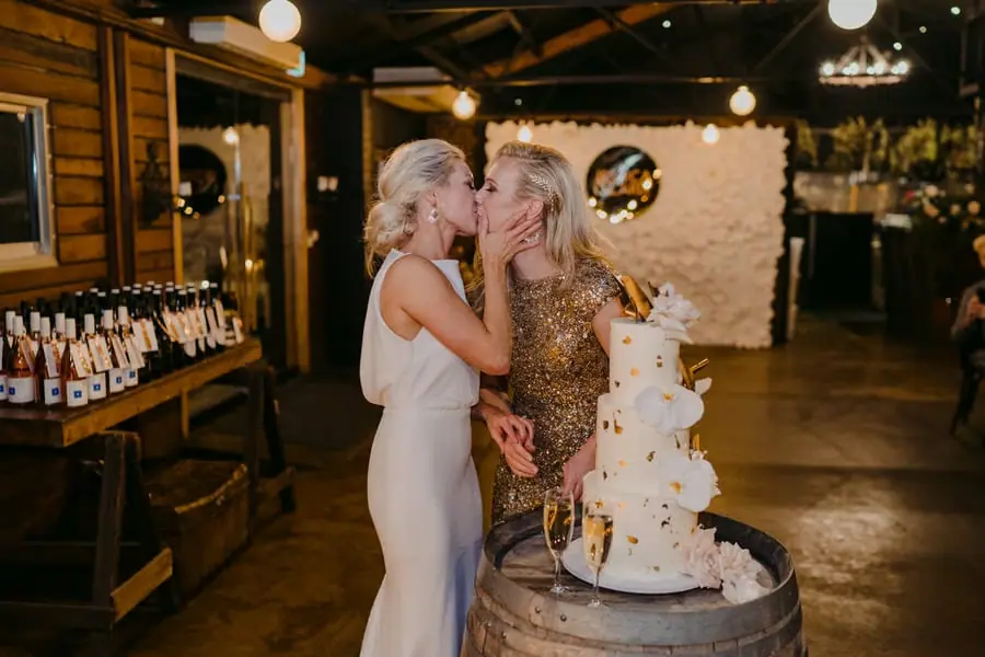 Two brides kissing beside a three-tier white wedding cake