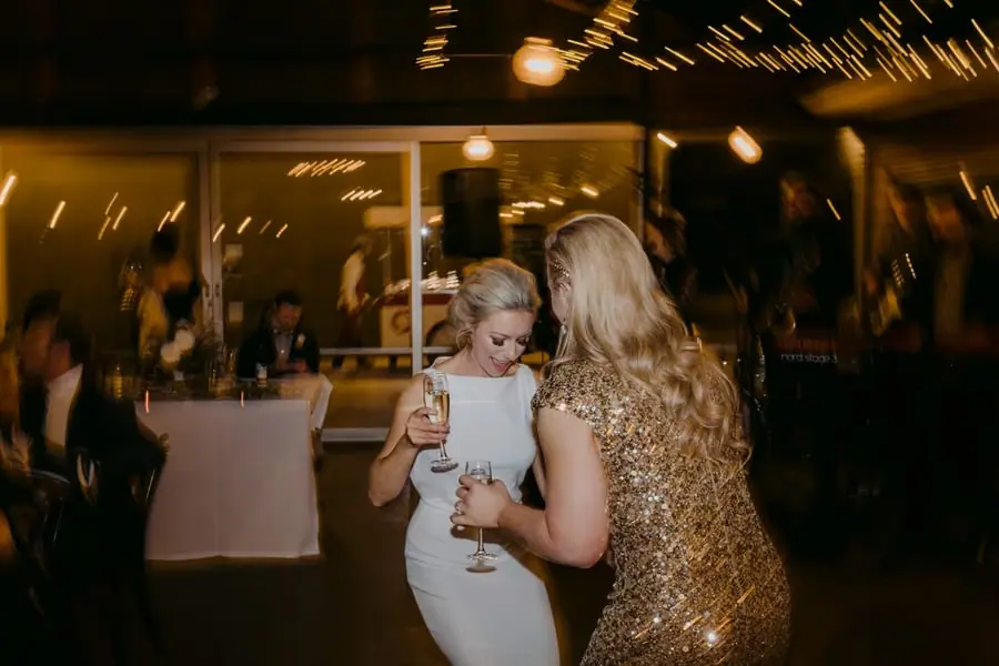 Two brides laughing with drinks beneath streaks of warm party lights