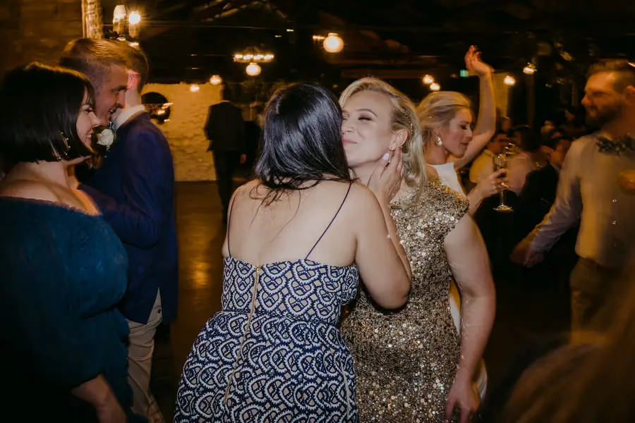 Bride in a gold dress embracing a smiling guest among the reception crowd