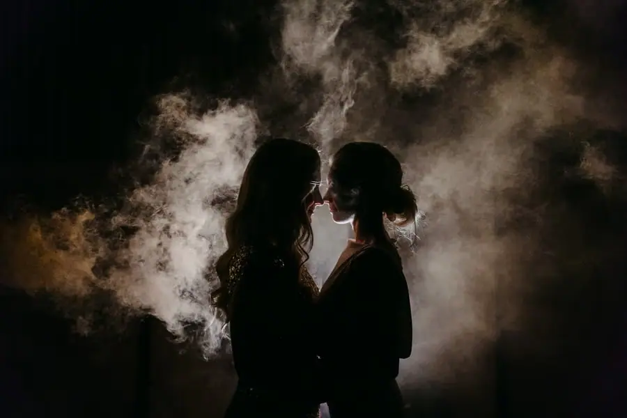 Silhouettes of two brides leaning close together against illuminated mist at night