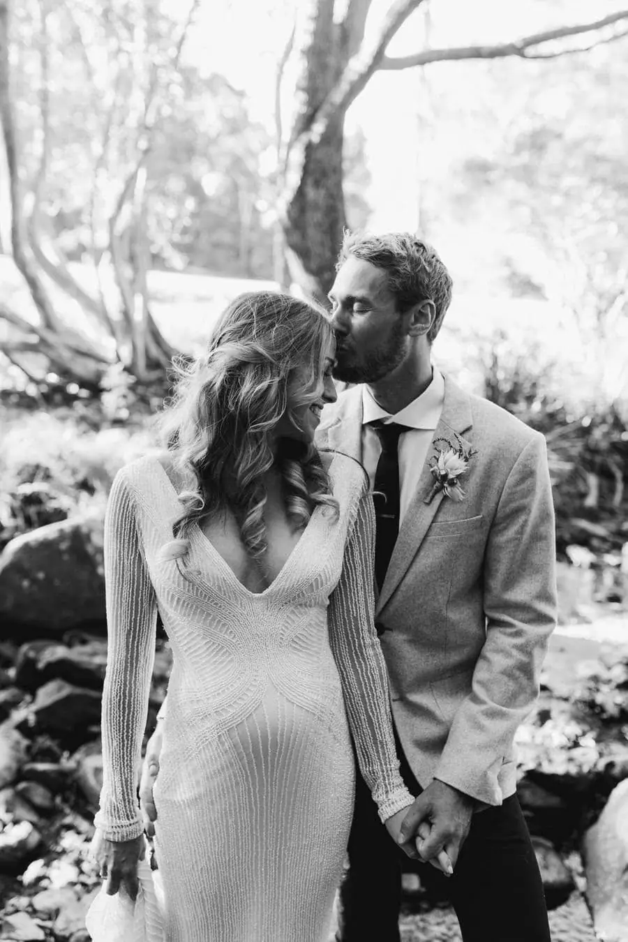 Black-and-white photograph of the groom kissing the bride beside a creek
