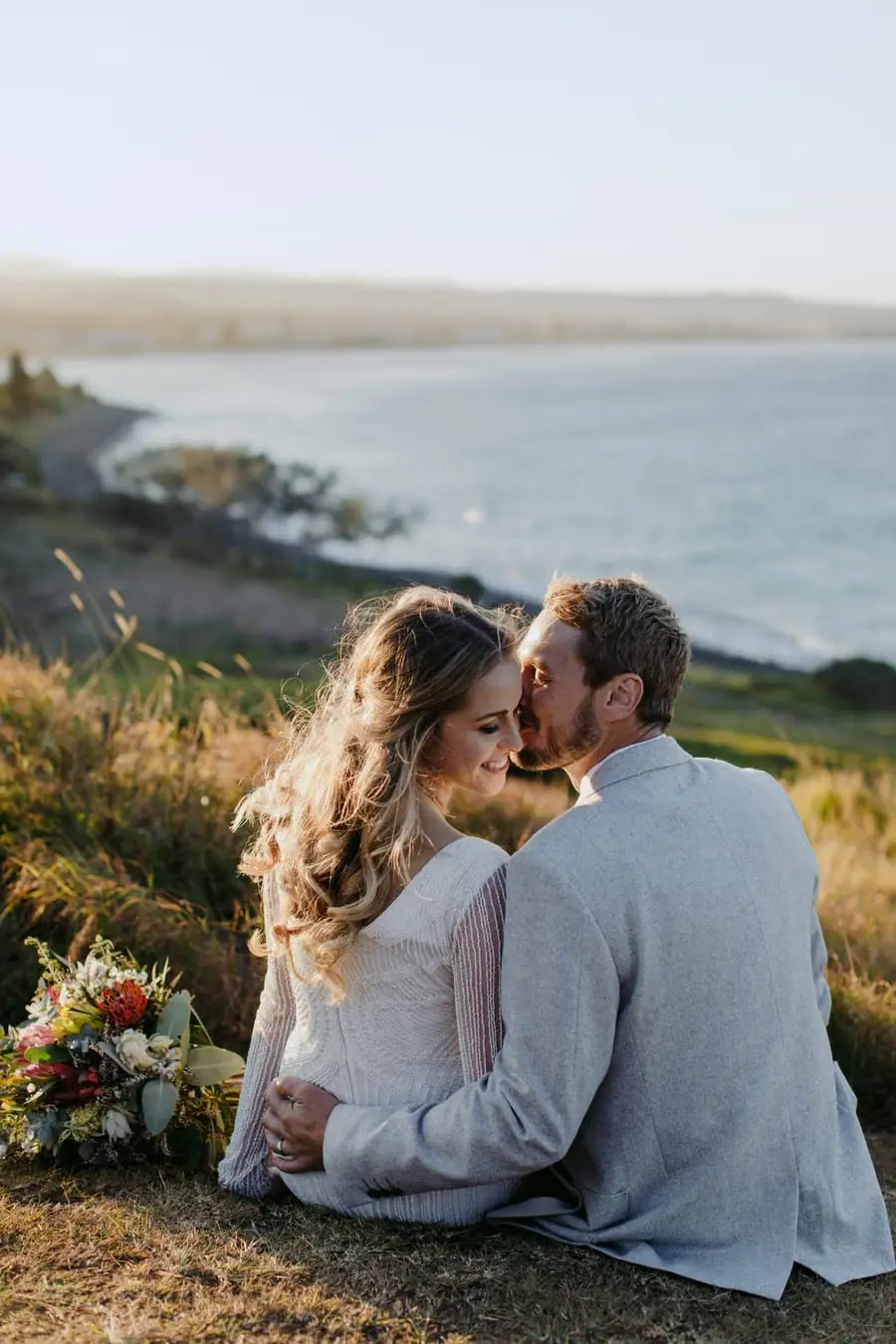 Newlyweds seated together on grass overlooking the rocky coastline