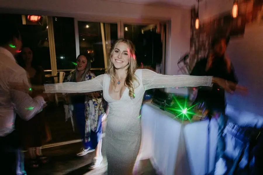 Bride dancing with her arms outstretched beside coloured party lights