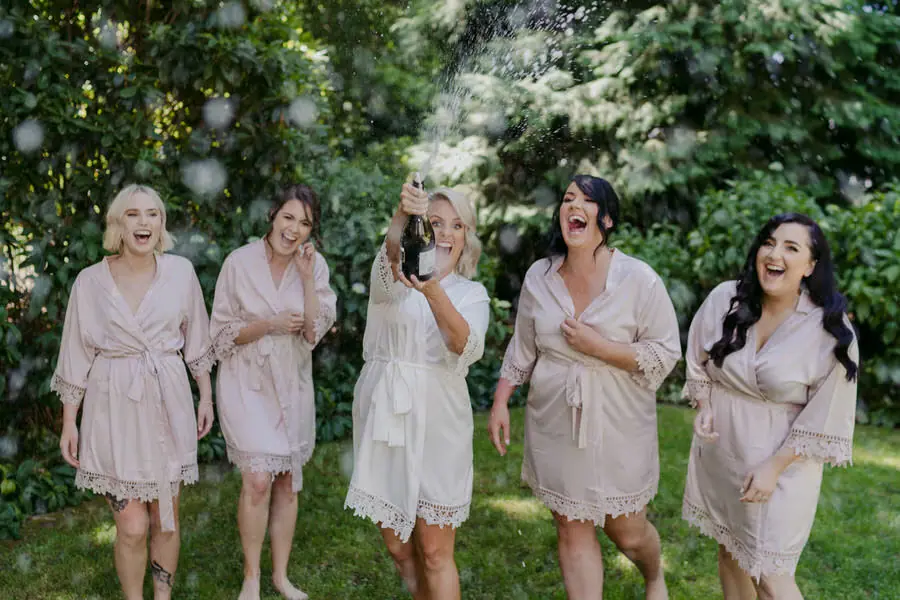 Bride opening a champagne bottle as friends laugh in the garden