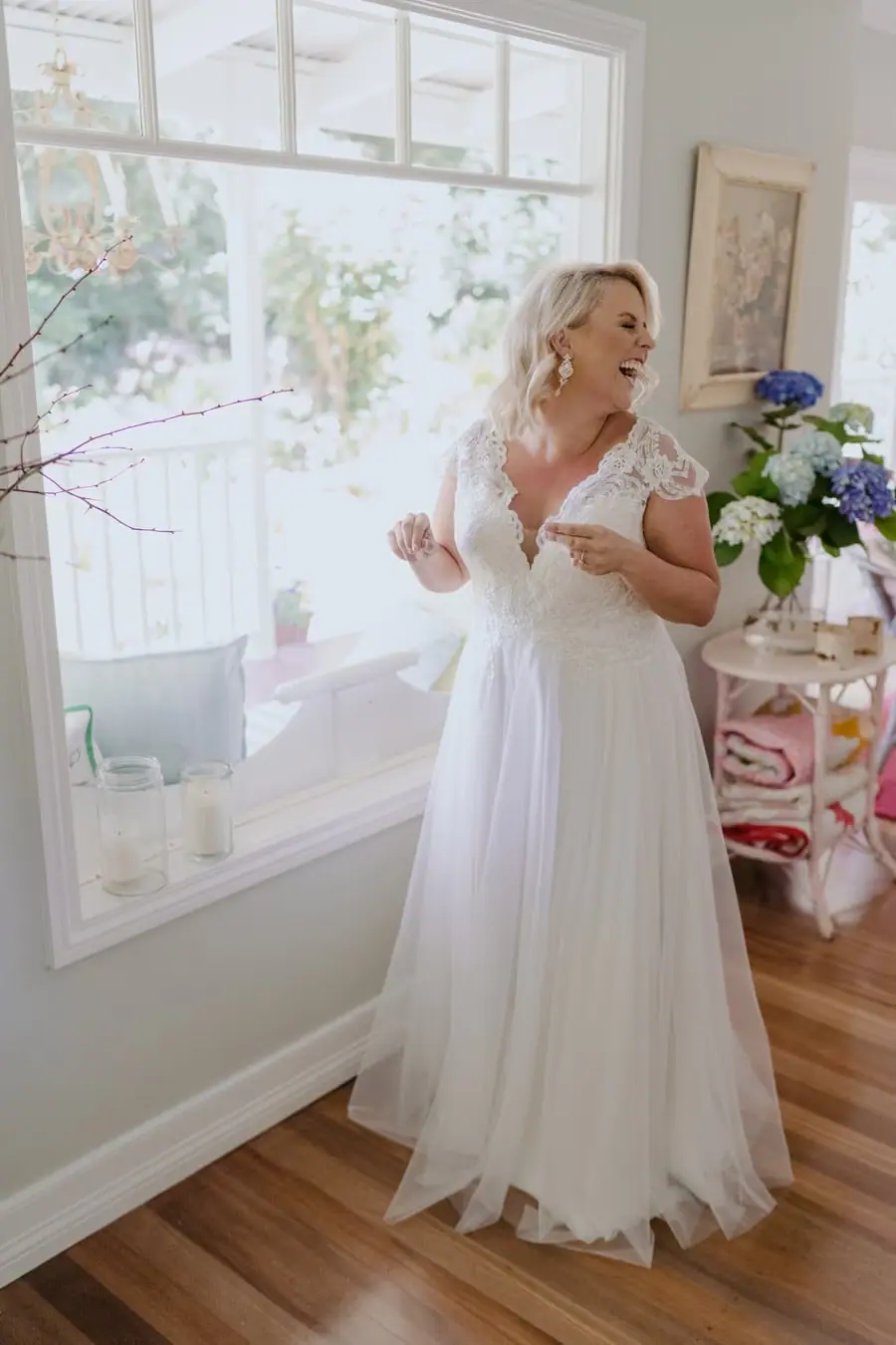 Bride smiling in her lace wedding dress beside a bright window