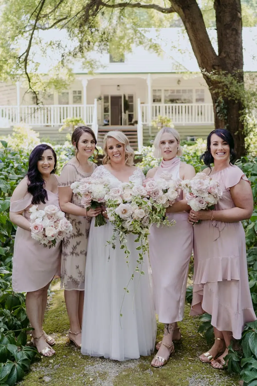 Bride and four friends posing with pale bouquets beneath a garden tree