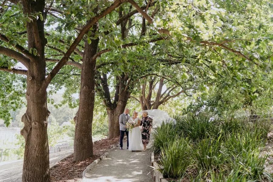 Bride walking with two companions along a tree-shaded path
