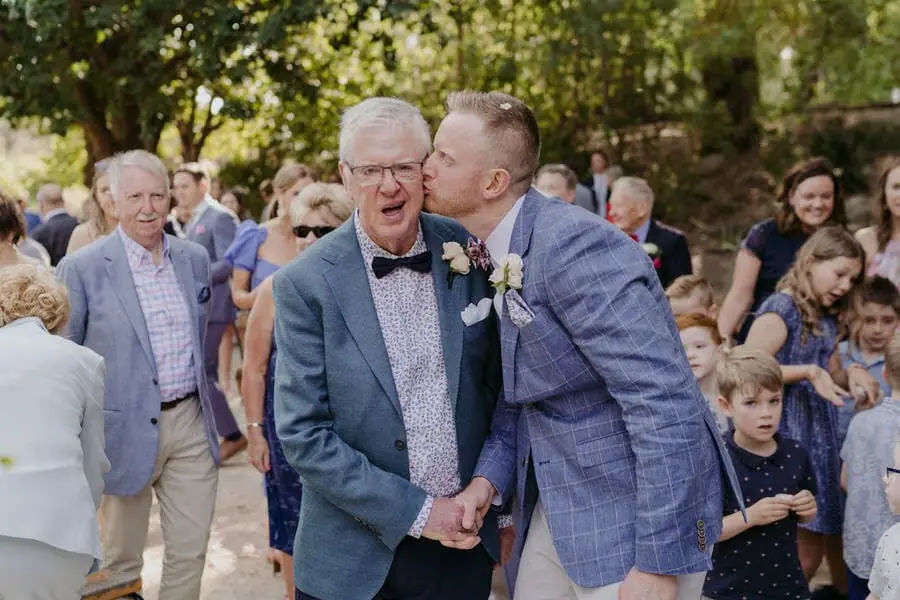 Groom kissing an older man's cheek among gathered wedding guests