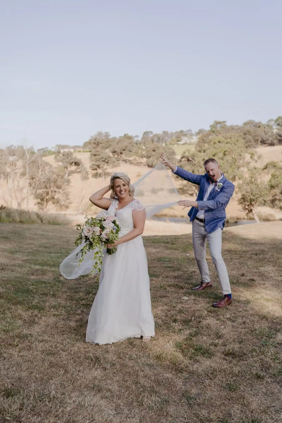 Groom raising an arm playfully as the bride smiles beside him outdoors