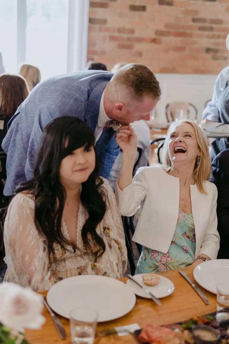 Groom leaning toward a laughing guest at the reception table