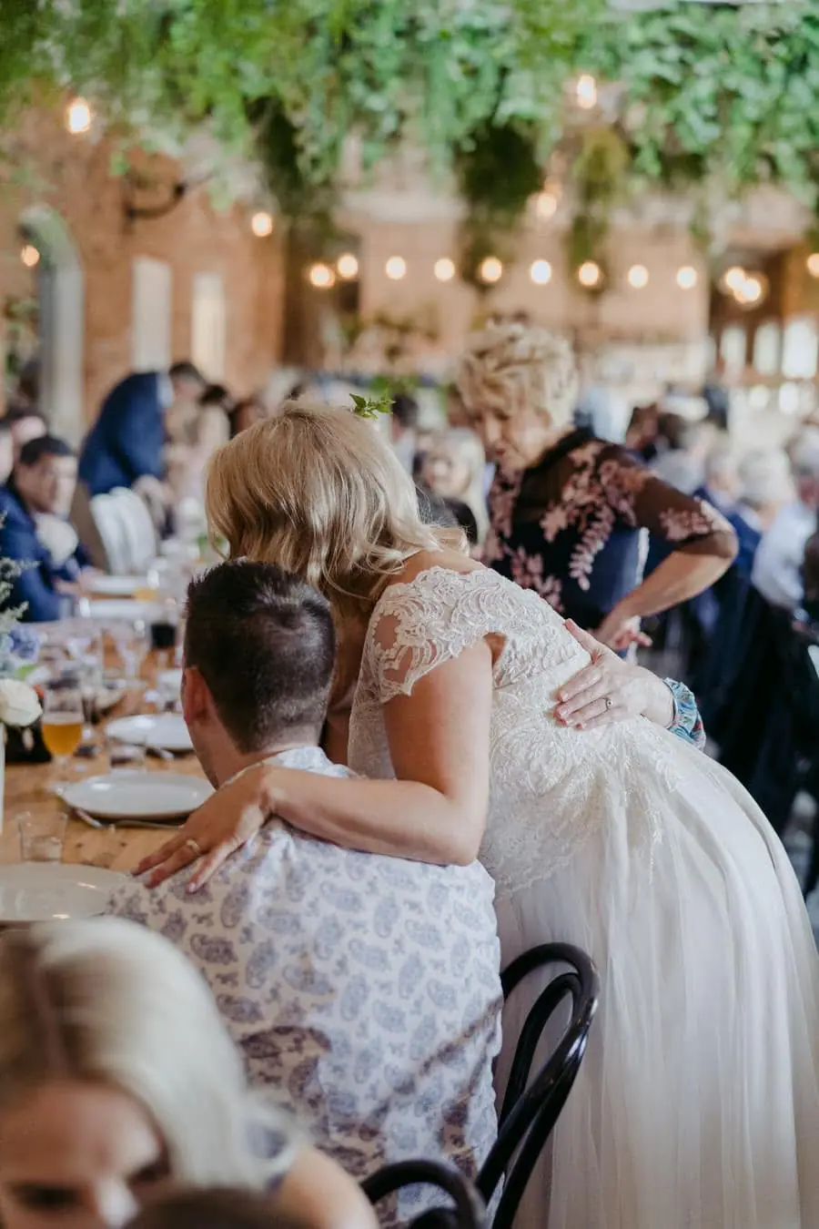 Bride embracing a seated guest across a wooden reception table