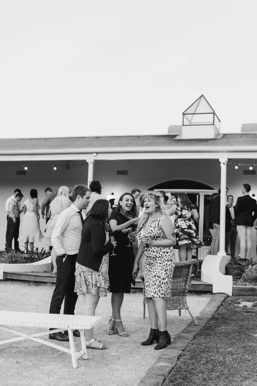Black-and-white photograph of guests standing with drinks near the verandah