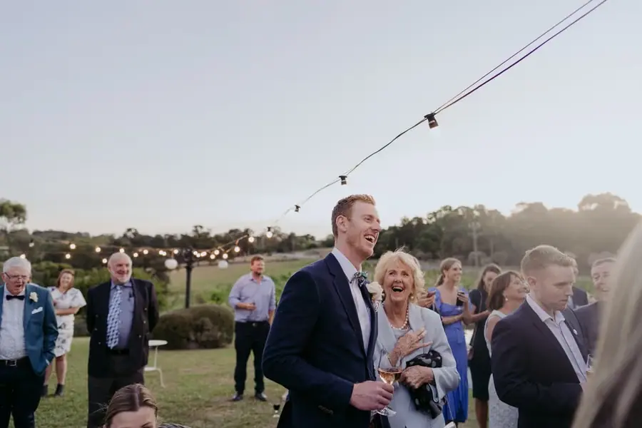 Bride and groom laughing beside standing guests beneath festoon lights