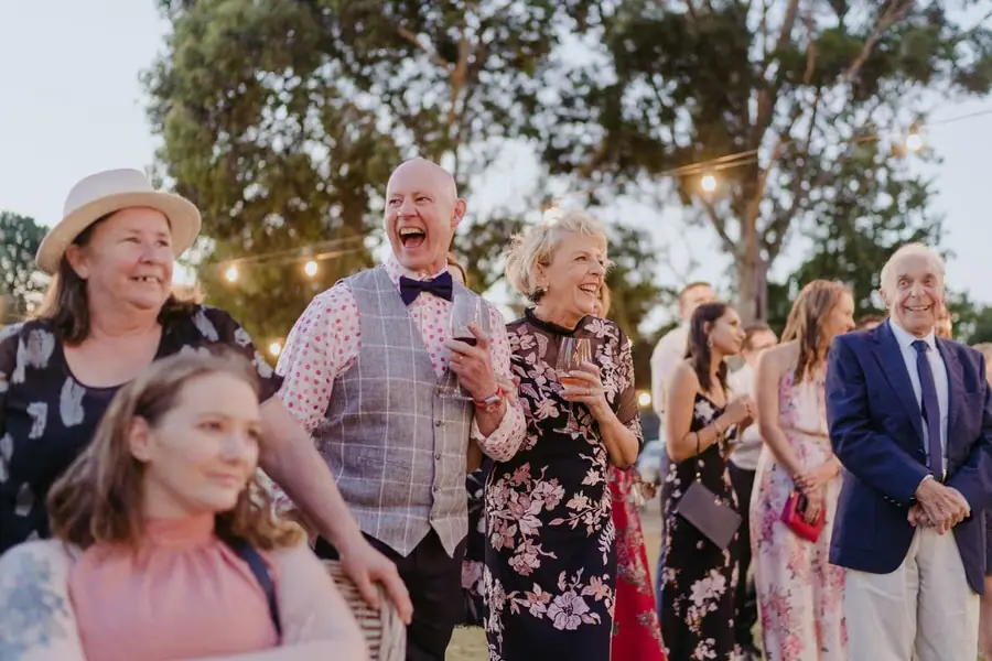 Older guests laughing together with drinks beneath garden trees