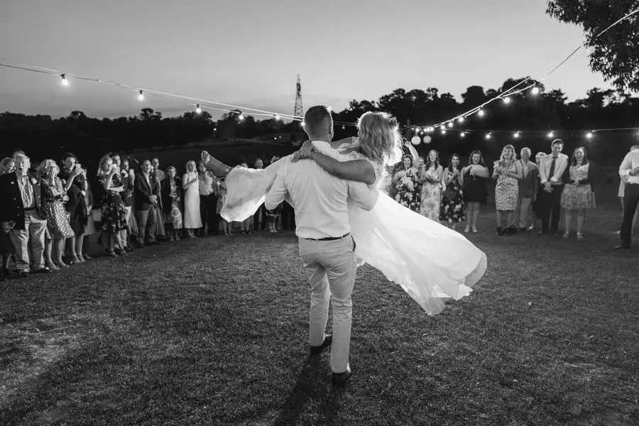 Black-and-white photograph of the groom carrying the bride as guests watch