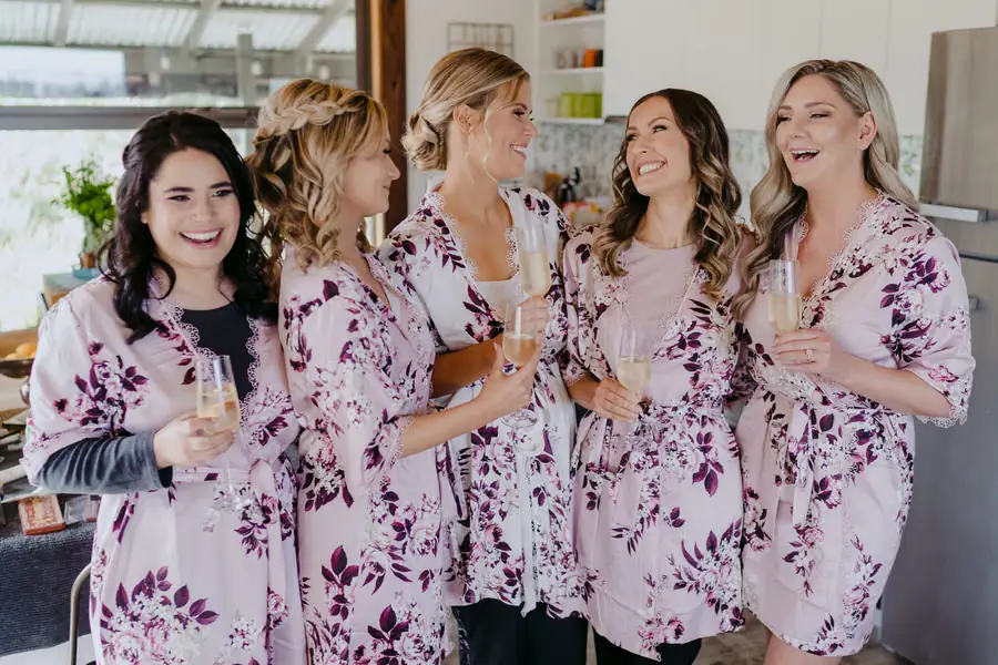 Bride and bridesmaids laughing together in matching floral robes with glasses