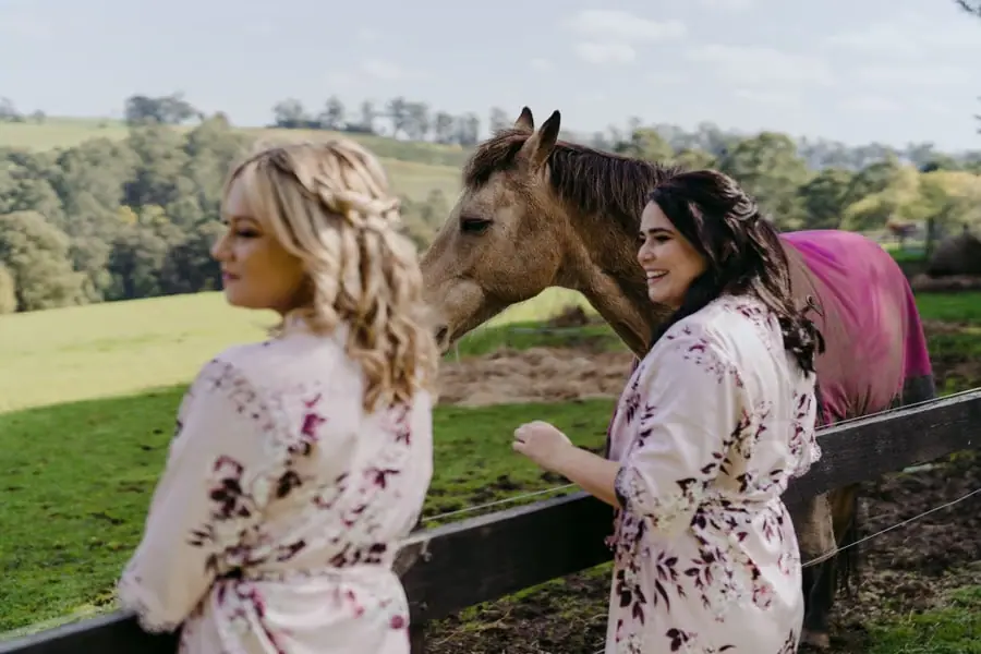 Two women in floral robes greeting a horse beside a paddock fence