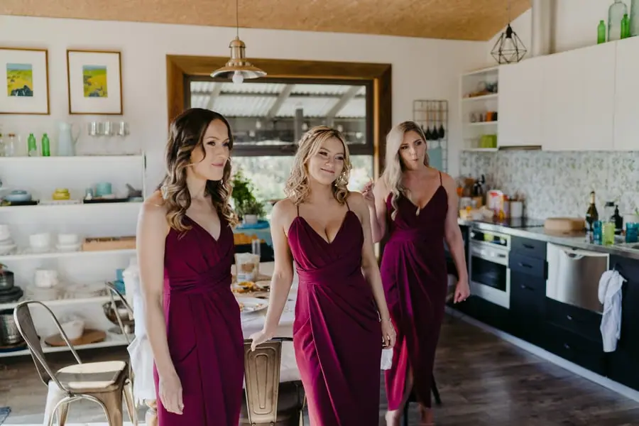 Three bridesmaids in burgundy dresses standing together in a kitchen