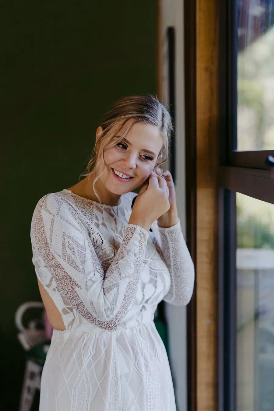 Bride smiling while putting on an earring beside a window