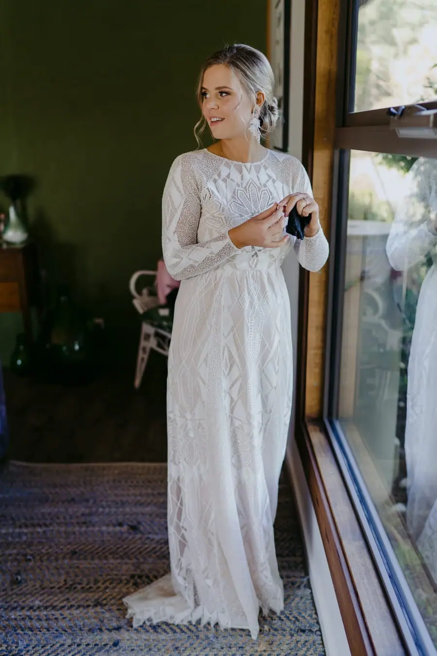 Bride standing beside a window with her hands clasped in her lace dress