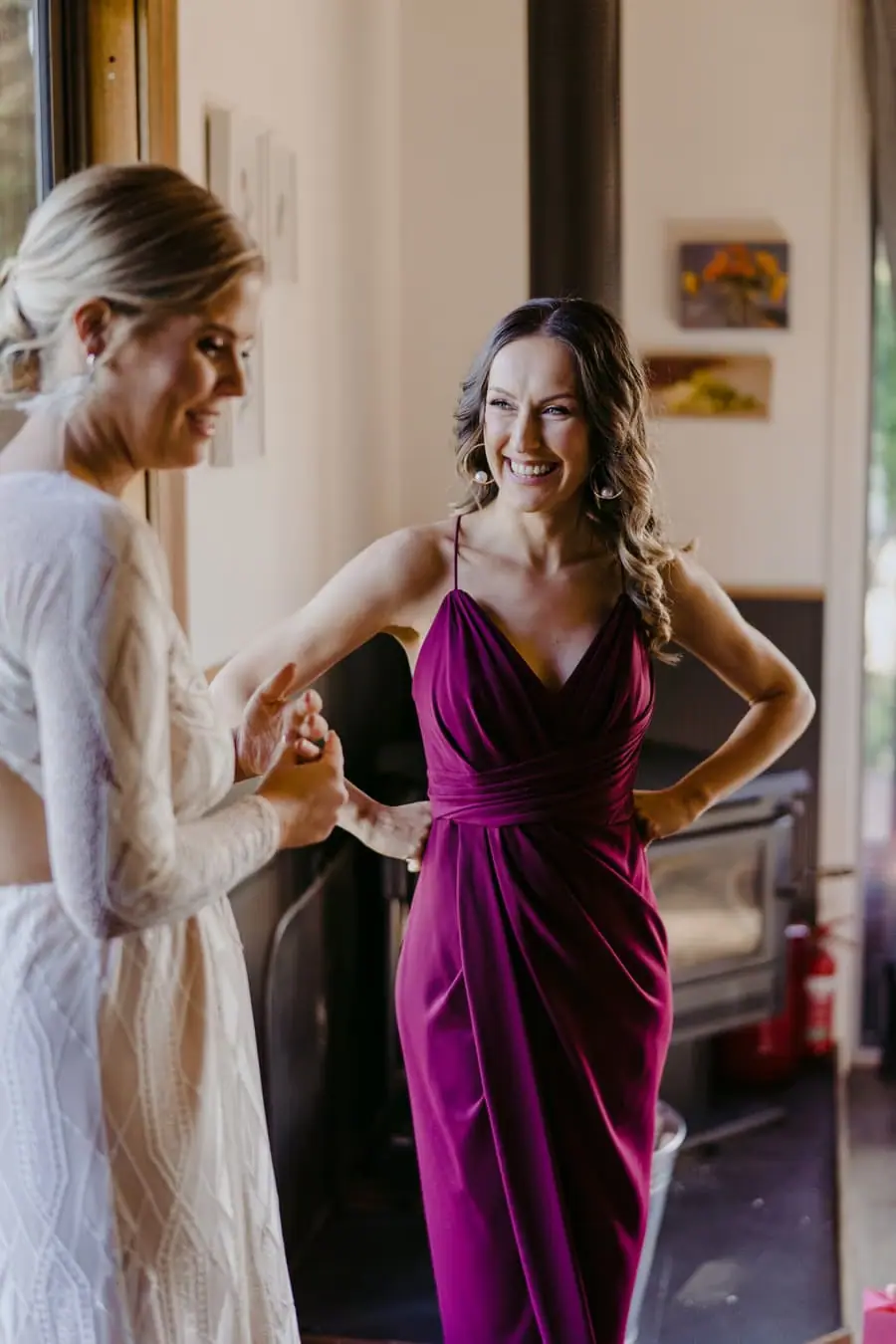 Bride and bridesmaid smiling together while holding drinks indoors