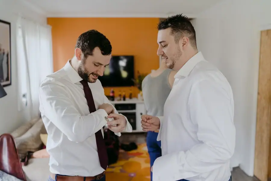 Two men smiling while adjusting shirt cuffs indoors