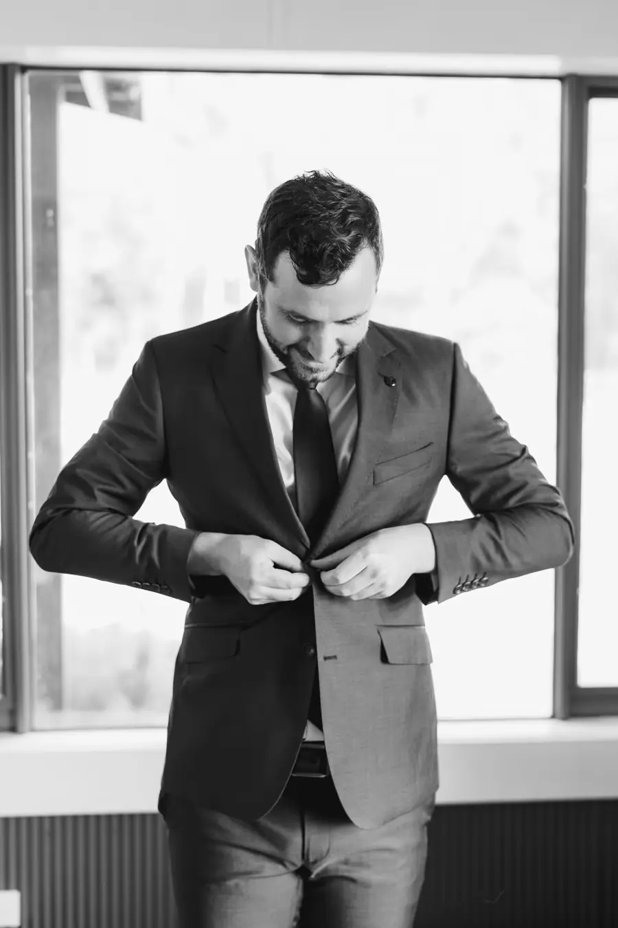 Black and white photograph of the groom fastening his suit jacket