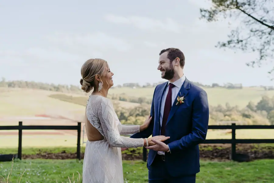 Bride and groom smiling at each other and holding hands beside a fence