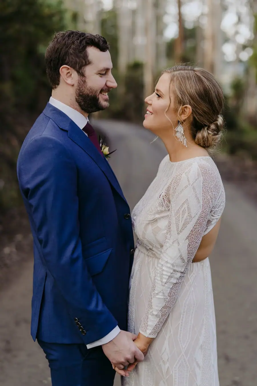 Bride and groom smiling at each other while holding hands on a forest road
