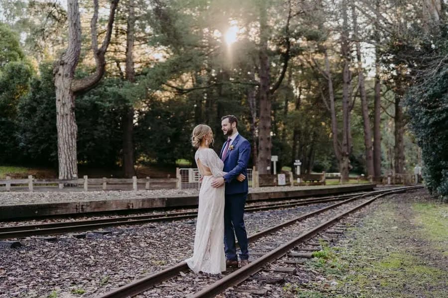 Groom embracing the bride from behind on railway tracks beneath trees