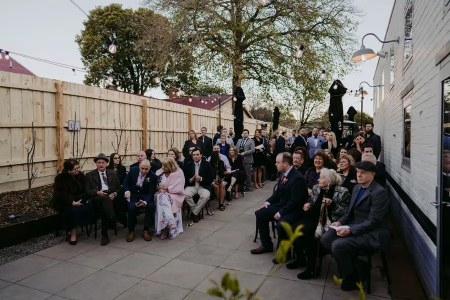 Wedding guests seated in a fenced courtyard beneath festoon lights