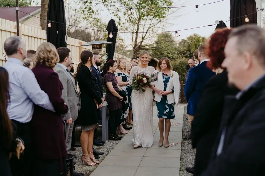 Bride entering the courtyard ceremony beside a woman as guests stand