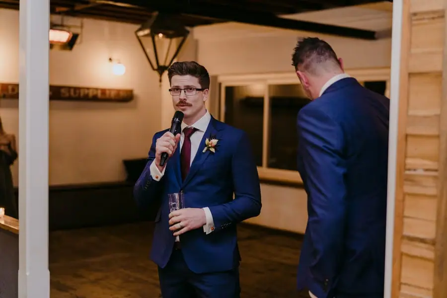 Groomsman with glasses speaking into a microphone beside another groomsman