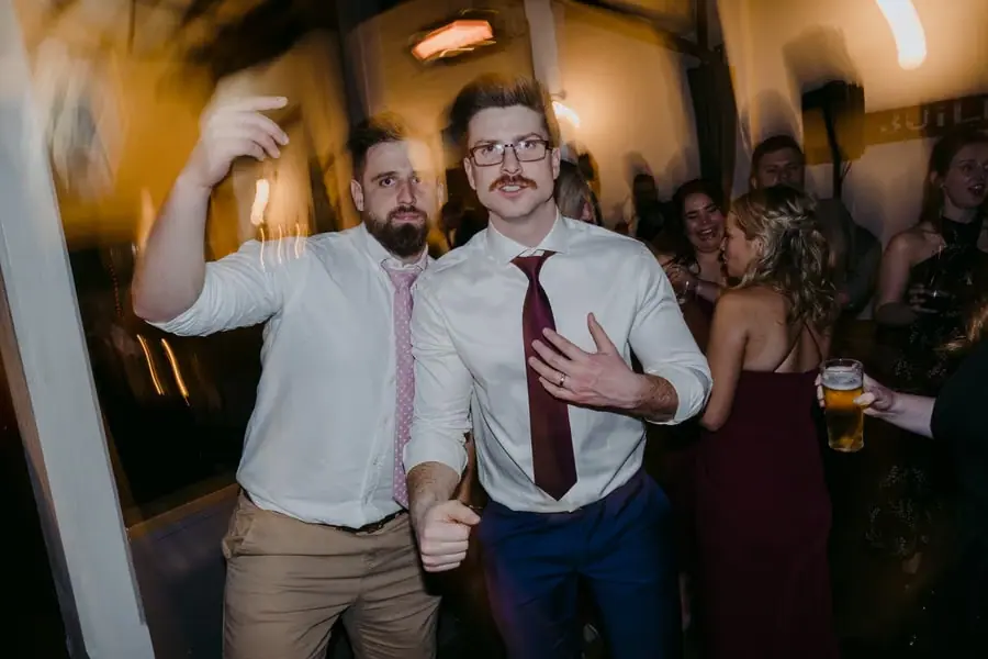 Men smiling and gesturing while dancing among reception guests