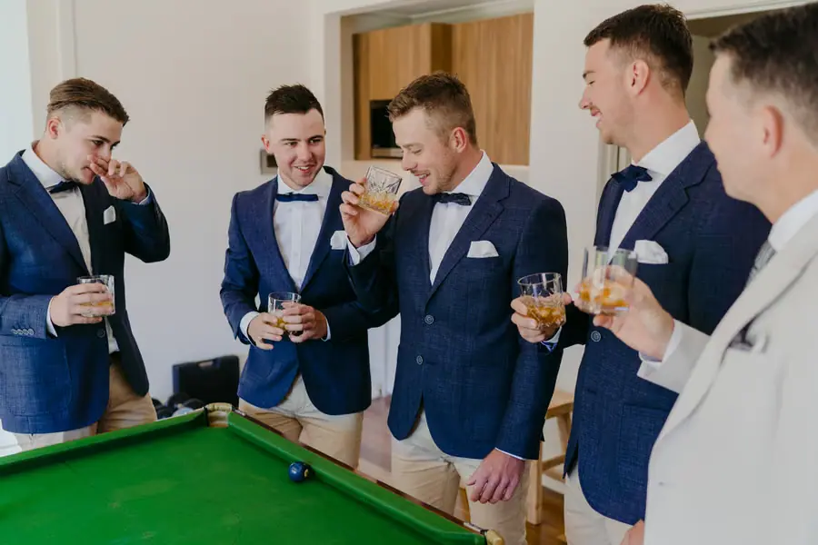 Groomsmen in blue jackets laughing with drinks beside a pool table