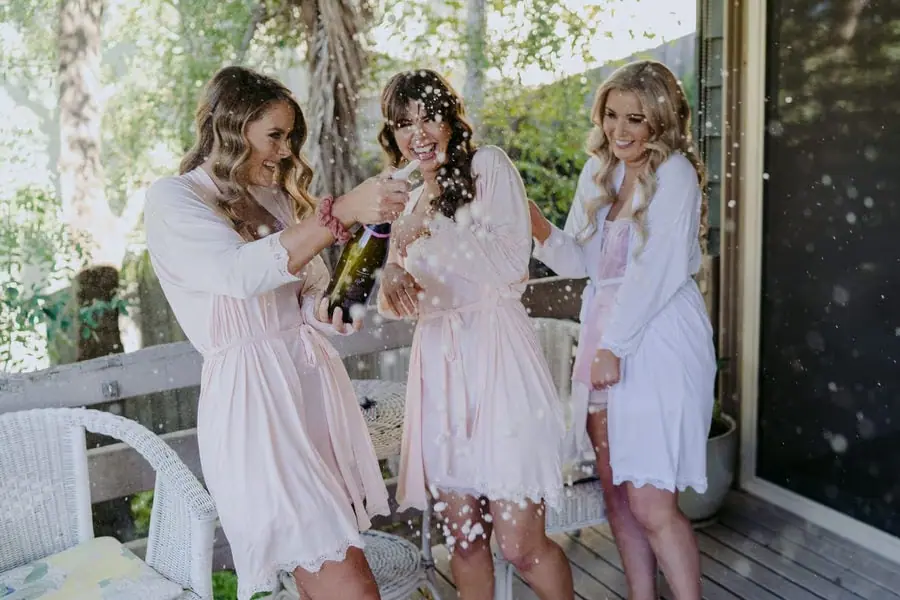 Women in pale robes laughing as sparkling wine sprays on a balcony
