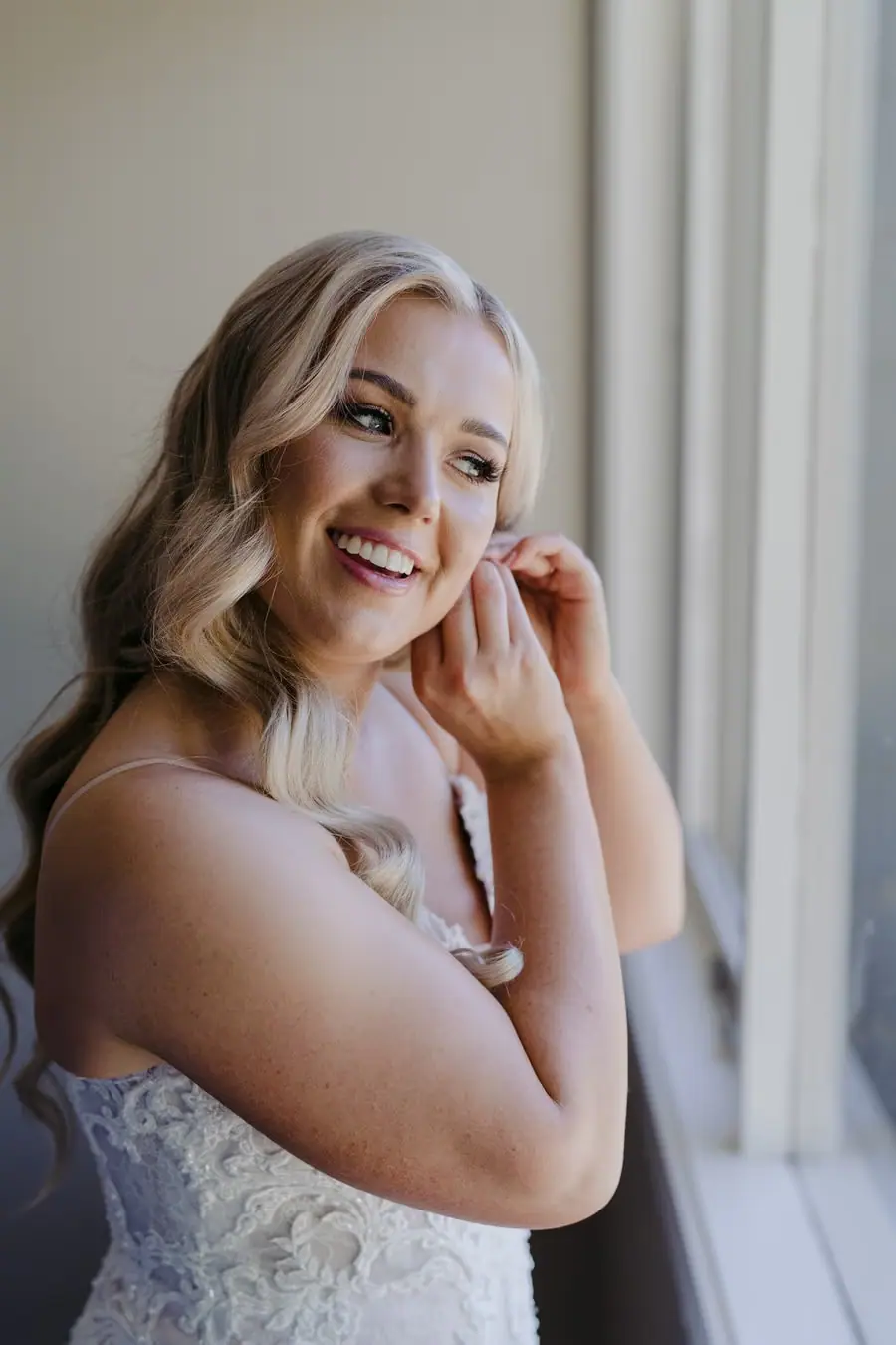 Bride smiling while fastening an earring beside a window
